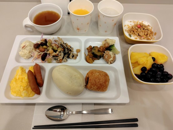 メインのお皿：左上からポテトサラダ、豆、蓮根とひじきのサラダ、鯖と茄子のボロネーゼ、おから肉団子とブロッコリー
パンオショコラ、白いパン、小さめのウィンナーとスクランブルエッグ
お皿回り：左からオニオンスープ、紙カップに入れられたスムージー、グラノーラ載せたヨーグルト、パイナップルとブルーベリー